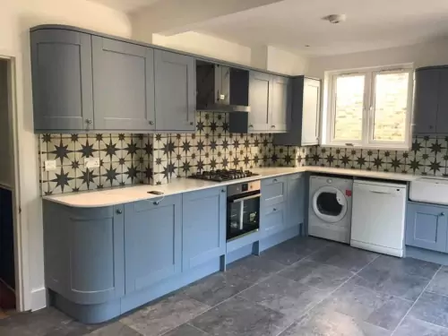 Modern L-shaped kitchen with light blue cabinets, patterned tile backsplash, built-in oven and stovetop, washing machine and dishwasher, gray floor tiles, and a window letting in natural light.